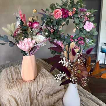 Two elegant vases with dried floral arrangements on a textured cloth and a floral wreath in the background.