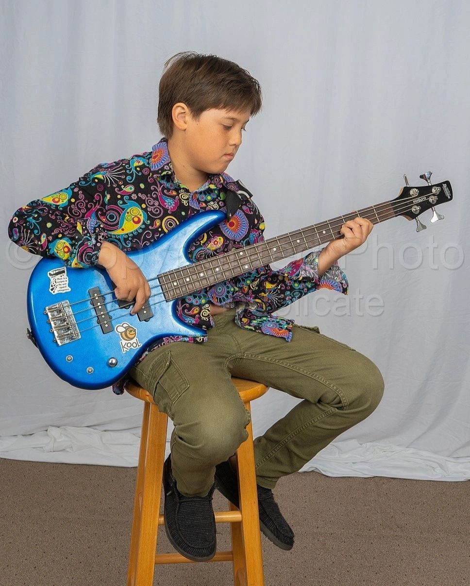 Young boy playing a blue electric bass guitar while sitting on a wooden stool.
