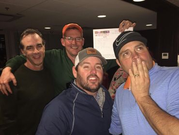 Four men posing indoors with one holding a paper.