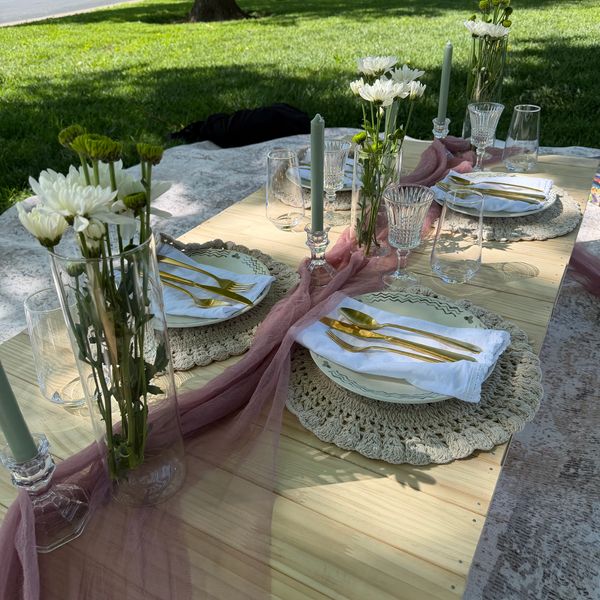 Elegant outdoor table setting with flowers, candles, and gold cutlery on a wooden table.