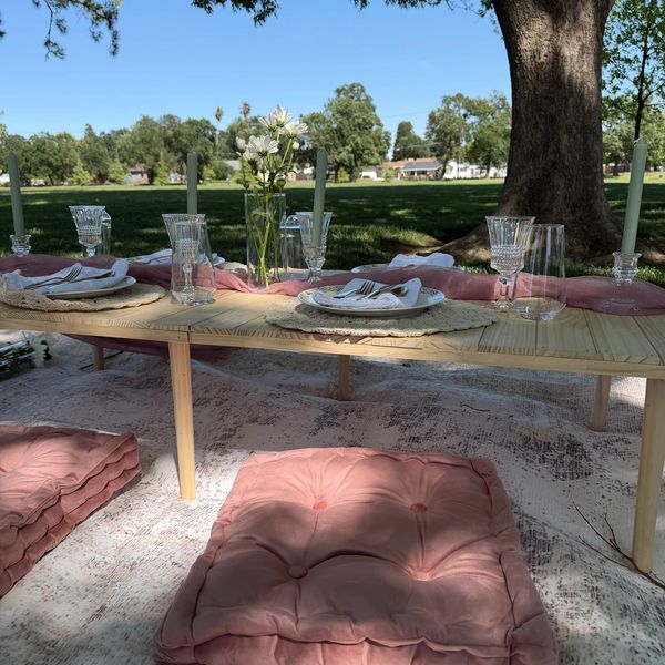 Outdoor picnic table set with pink cushions, flowers, and candles under a large tree.