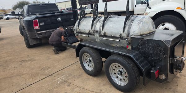 A man working on a trailer-mounted barbecue smoker attached to a black pickup truck.