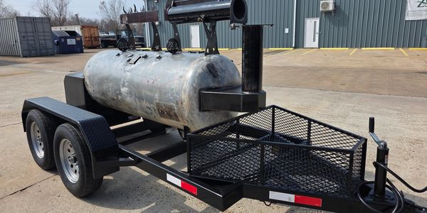 A large metal smoker grill mounted on a dual-axle trailer in an industrial area.