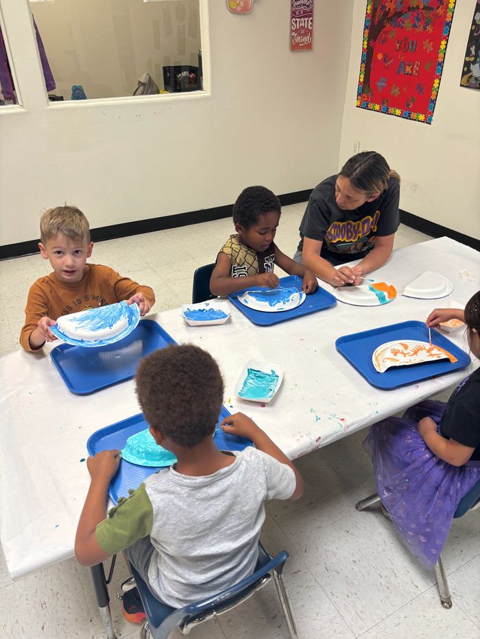 Kids and adult painting on paper plates in classroom.