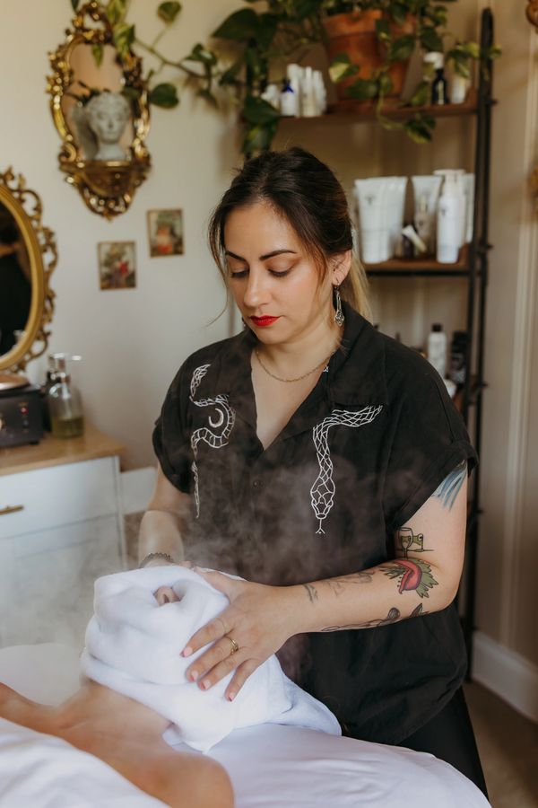 Woman performing a hot towel facial treatment in a cozy room.
