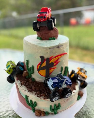 Two-tier monster truck themed cake with toy trucks and cacti decorations.