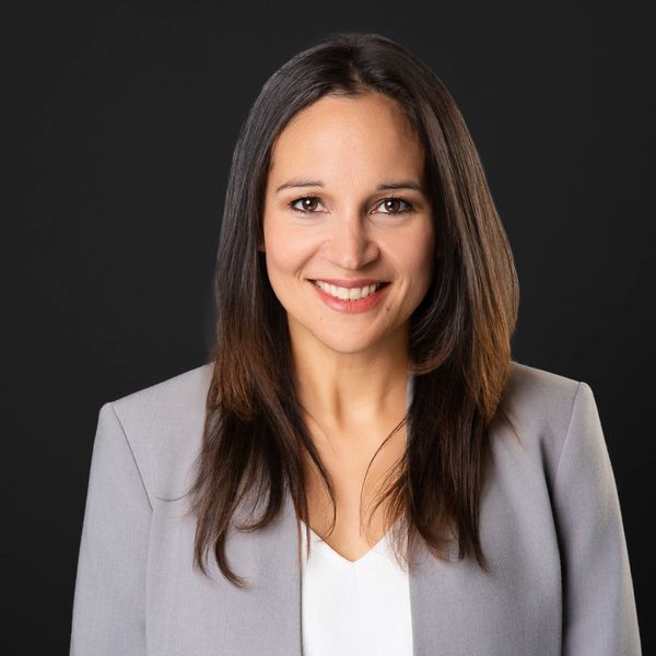 Smiling professional woman in a gray blazer against black background.