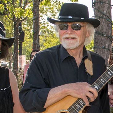 Elderly man in black hat and sunglasses playing guitar outdoors.