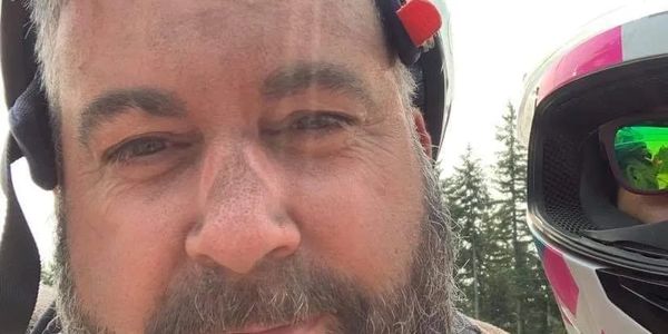 Man with a beard lifting a green helmet outdoors.