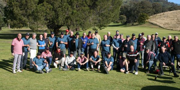 Large group of people posing outdoors on a sunny day, some seated on grass.