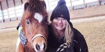 Woman smiling next to a brown horse indoors.