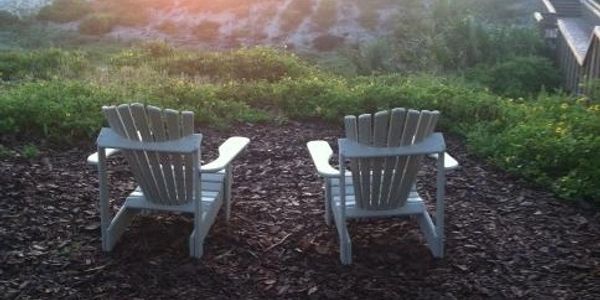 Two white Adirondack chairs on mulch facing a garden at sunset.