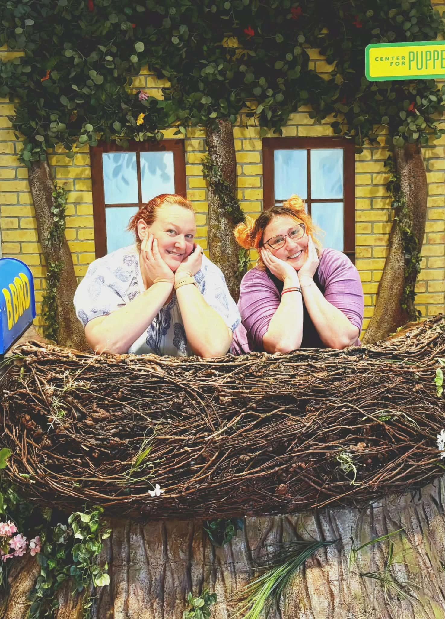 Two women smiling, leaning on a large bird nest backdrop.