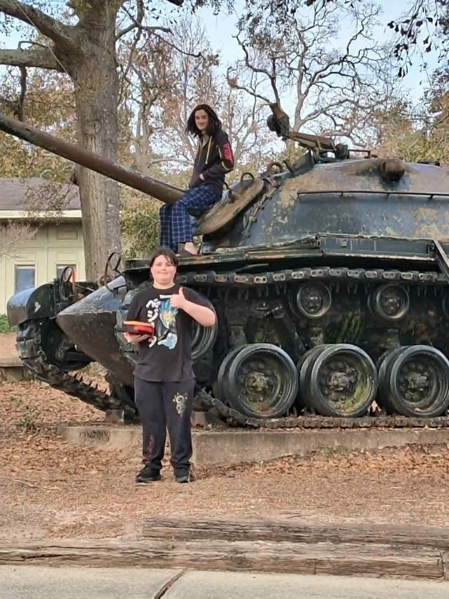 Two kids posing with an old tank outdoors in a park-like setting.
