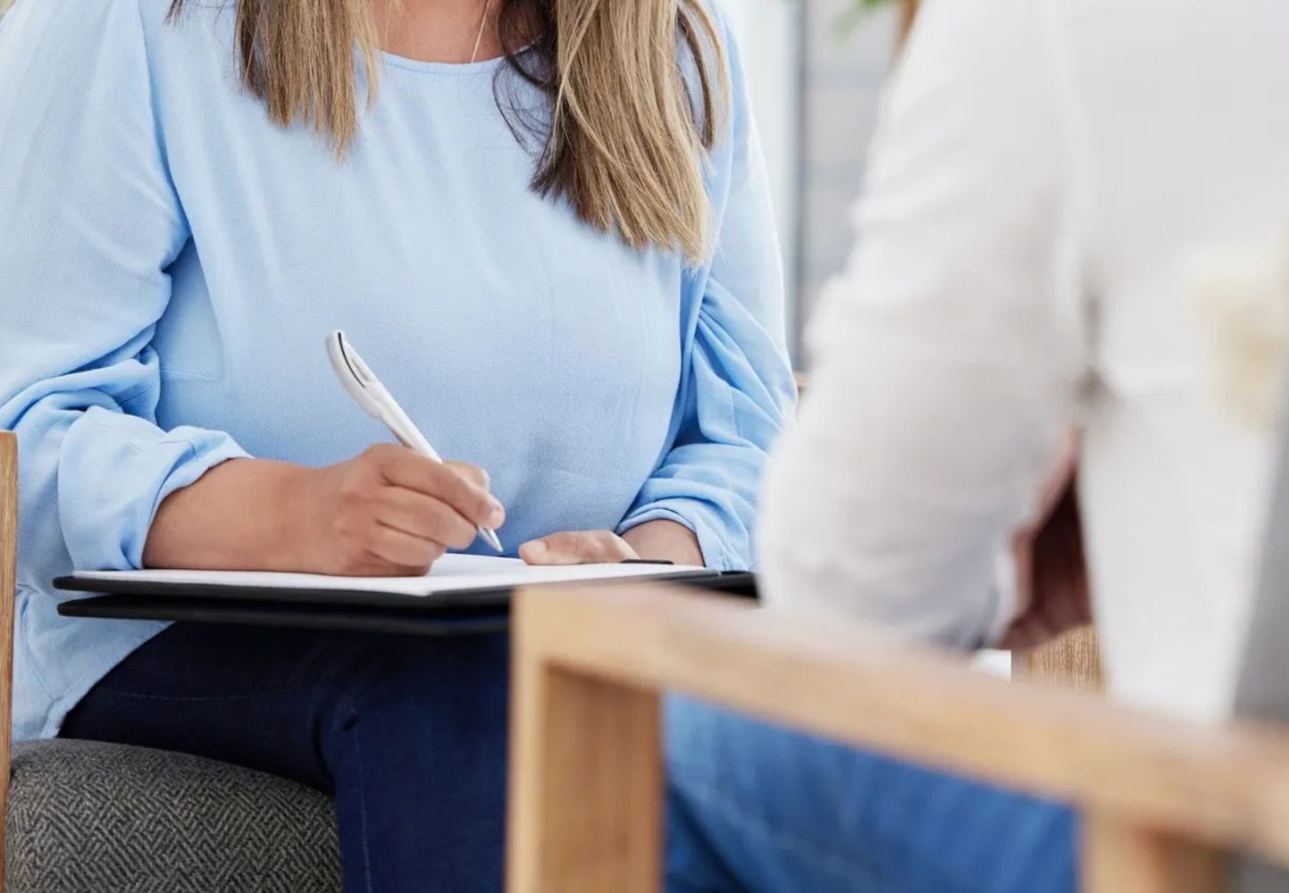 Woman in blue blouse writing on a clipboard during a conversation.