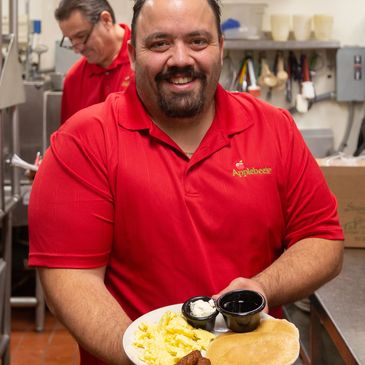 Smiling Applebee's manager holding a breakfast plate with pancakes, scrambled eggs, and sausage.