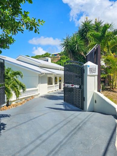 Entrance gate to a bright white villa surrounded by tropical greenery under a clear blue sky.