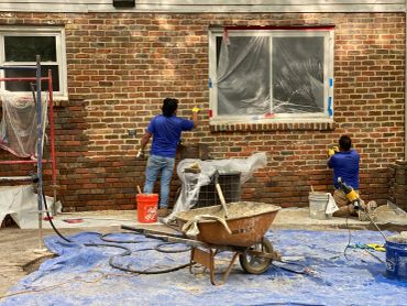 Two workers repairing a brick wall outside a house.