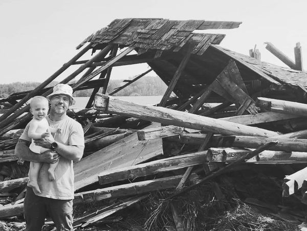 Man holding baby in front of collapsed wooden structure.