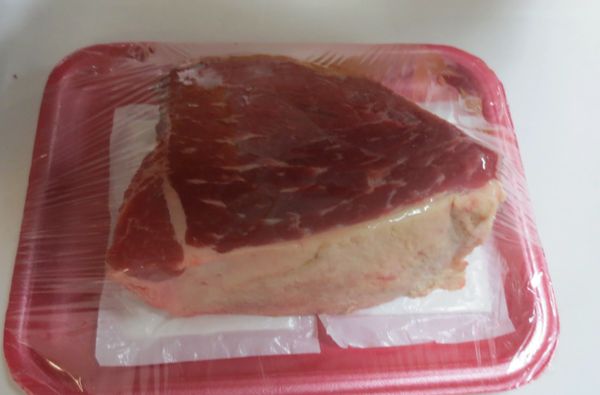 A thick piece of raw meat wrapped in plastic on a red tray.