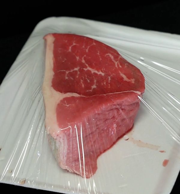 Raw piece of beef steak wrapped in plastic on a white tray.