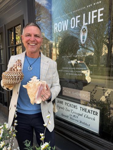 Man holding two large seashells in front of a theater poster for 'Row of Life'.