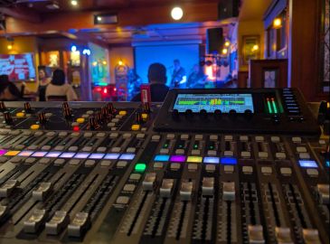 Sound mixer controls in a dimly lit live music venue with a band playing.