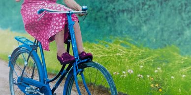 Elderly woman in pink dress and hat rides a blue bicycle, waving joyfully with birthday wishes.