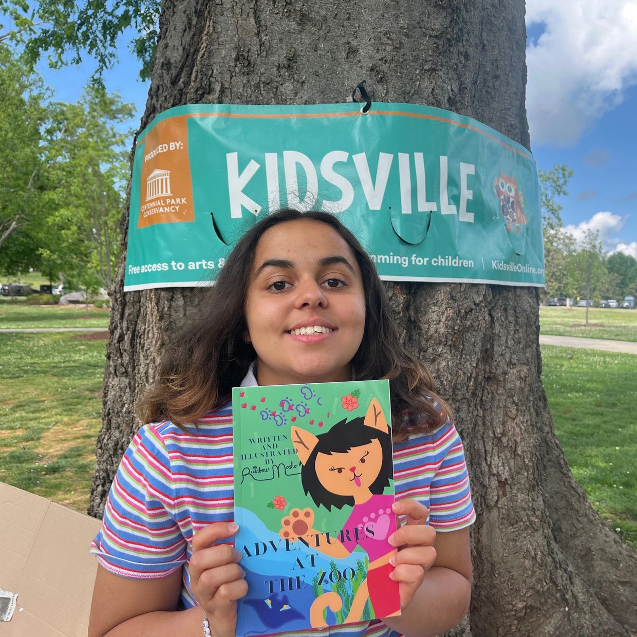 A girl holding a colorful children's book in front of a Kidsville banner on a tree.