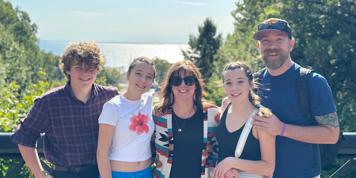 A smiling family of five posing outdoors on a sunny day with greenery and water in the background.