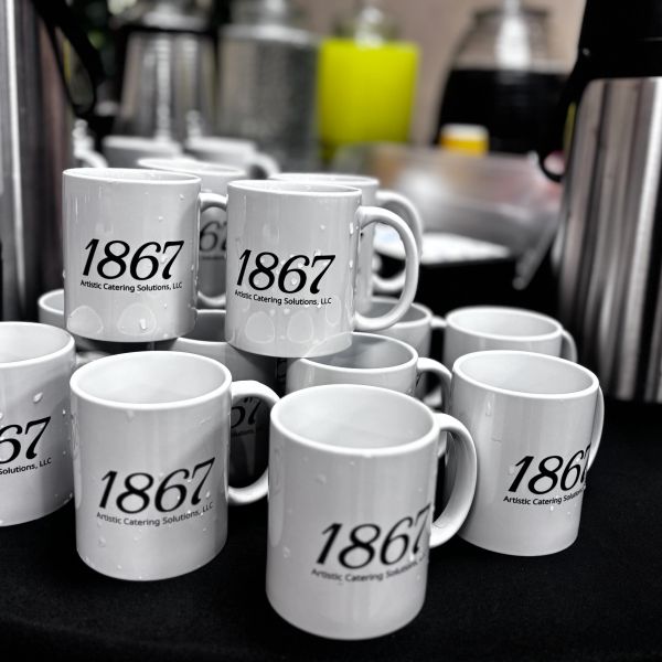 Stack of white mugs with '1867 Artistic Catering Solutions, LLC' printed on them.