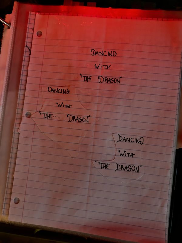 Handwritten phrases "Dancing with the Dragon" repeated on lined paper with red lighting.