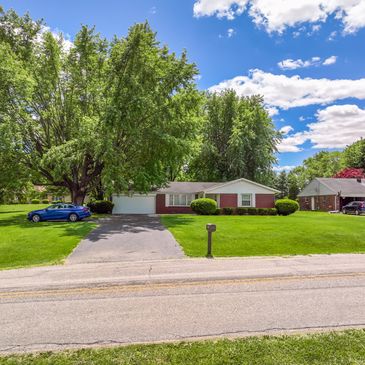 Single-story house with lush green lawn and large trees under a blue sky.