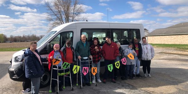 Group of people holding a colorful "Thank You" banner in front of a white van.
