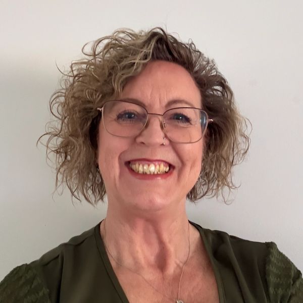 Smiling woman with curly hair and glasses wearing a green top and heart necklace.