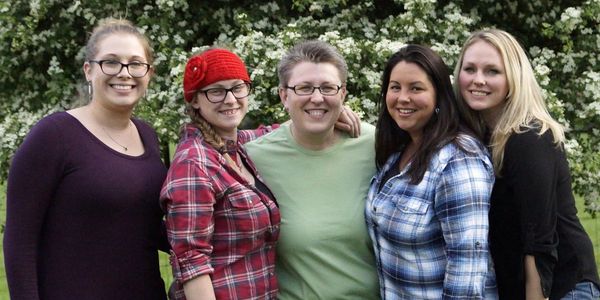 Five women smiling outdoors with flowering bushes behind them.