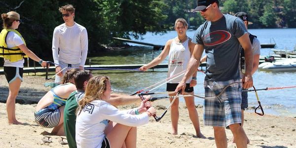 Group learning slingshot use on a lakeside beach with instructor.