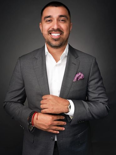Smiling man in charcoal suit and white shirt, purple pocket square, studio headshot