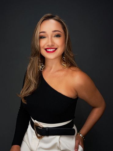 Smiling woman in black one-shoulder top and white pants, gold earrings, studio headshot