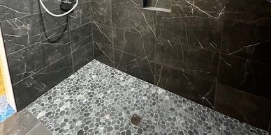 A modern shower with black marble walls and pebble stone floor.