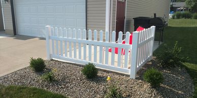 A section of a white picket fence on a gravel bed in front of a residential home.