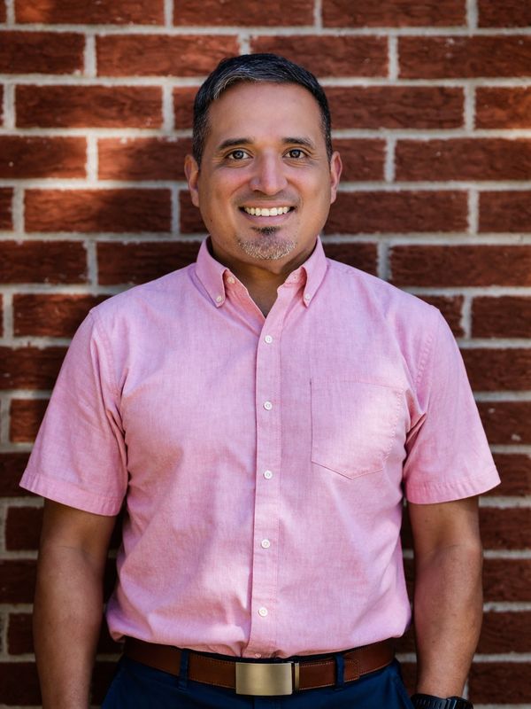 Man smiling in a pink shirt against a brick wall.