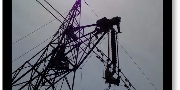 Workers climbing and repairing a tall electricity transmission tower against a cloudy sky.