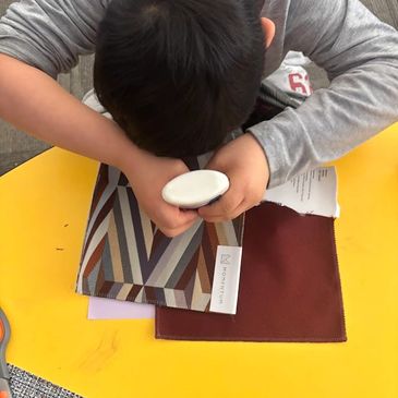 Child closely examines a fabric sample with a magnifying glass on a yellow table.