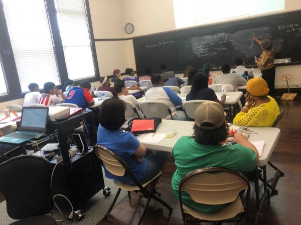 A classroom with students attentively listening to a teacher writing on a chalkboard.