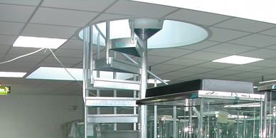 A galvanised steel spiral staircase serving an office mezzanine floor through a circular void.