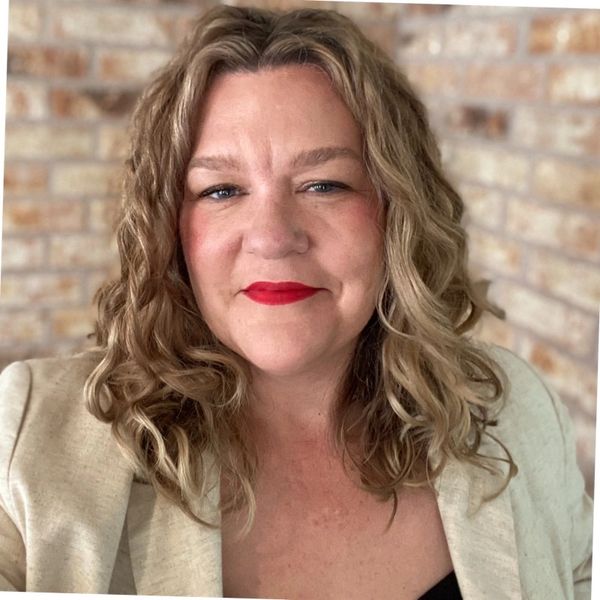 Smiling woman with curly hair and red lipstick against a brick wall.
