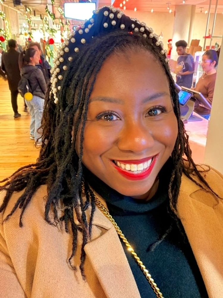 #BeingSRosette is a woman pictured smiling while wearing a pearl headband and camel cashmere coat.