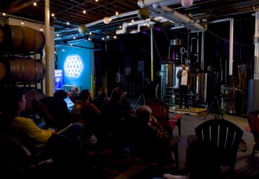 A comedian performs on stage in a cozy brewery with an audience seated in Adirondack chairs.