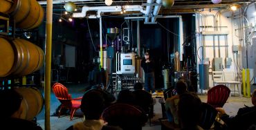 Audience watching a speaker in an industrial-style room with barrels and red chairs.